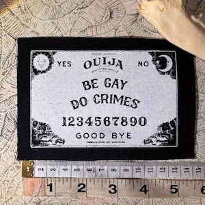 Op de afbeelding: Zwarte Ouija bord patch met de tekst "BE GAY DO CRIMES". De patch bevat de woorden "YES" en "NO", cijfers en de zin "GOOD BYE". De patch is ongeveer 13 cm lang.