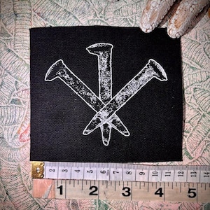 May include: Black square patch featuring a white illustration of three nails in a symbol. The patch is approximately 7.6 cm wide. The background is a patterned fabric.