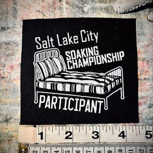 Può includere: Toppa in bianco e nero con il testo "Salt Lake City Soaking Championship Participant" e un'immagine di un letto con una testata a righe.