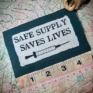 May include: A dark green patch with the text "SAFE SUPPLY SAVES LIVES" and a syringe graphic. The patch is approximately 10 cm long, measured with a ruler. The background has a patterned design.