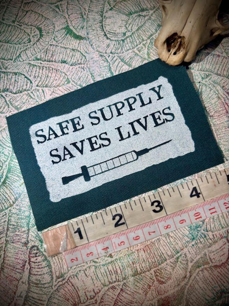 May include: A dark green patch with the text "SAFE SUPPLY SAVES LIVES" and a syringe graphic. The patch is approximately 10 cm long, measured with a ruler. The background has a patterned design.
