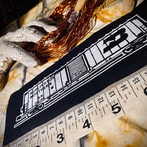 May include: A black patch featuring a white illustration of a Burlington Northern train car. The text "BURLINGTON NORTHERN" and "BN 355145" are visible. A measuring tape is placed near the patch. Copper wire and decorative elements are in the background.