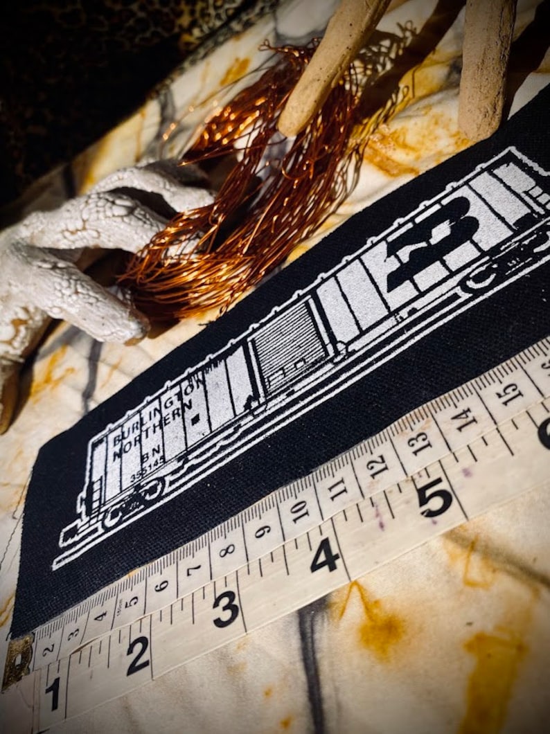 May include: A black patch featuring a white illustration of a Burlington Northern train car. The text "BURLINGTON NORTHERN" and "BN 355145" are visible. A measuring tape is placed near the patch. Copper wire and decorative elements are in the background.