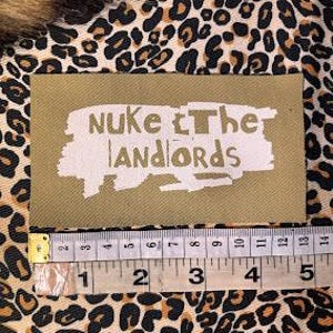 Peut inclure: Un &eacute;cusson rectangulaire vert olive avec le texte blanc "NUKE The LANDLORDS". Une r&egrave;gle en dessous montre les mesures en centim&egrave;tres. L'&eacute;cusson est sur un tissu &agrave; imprim&eacute; l&eacute;opard.