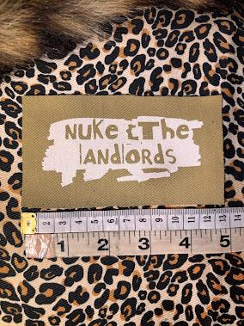 Peut inclure: Un &eacute;cusson rectangulaire vert olive avec le texte blanc "NUKE The LANDLORDS". Une r&egrave;gle en dessous montre les mesures en centim&egrave;tres. L'&eacute;cusson est sur un tissu &agrave; imprim&eacute; l&eacute;opard.