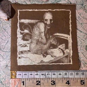 Puede incluir: Un parche de tela marr&oacute;n con una imagen en tonos sepia de una figura esquel&eacute;tica. La imagen est&aacute; enmarcada con un borde blanco. El parche mide aproximadamente 10 cm de ancho y 10 cm de alto.