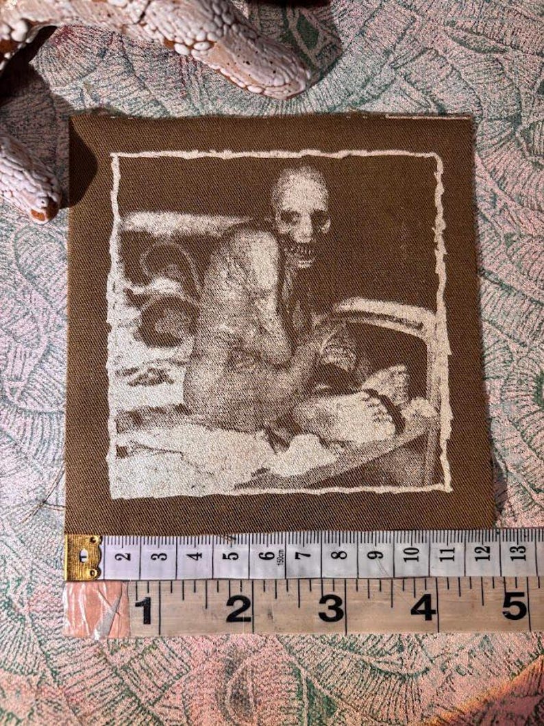 Puede incluir: Un parche de tela marr&oacute;n con una imagen en tonos sepia de una figura esquel&eacute;tica. La imagen est&aacute; enmarcada con un borde blanco. El parche mide aproximadamente 10 cm de ancho y 10 cm de alto.