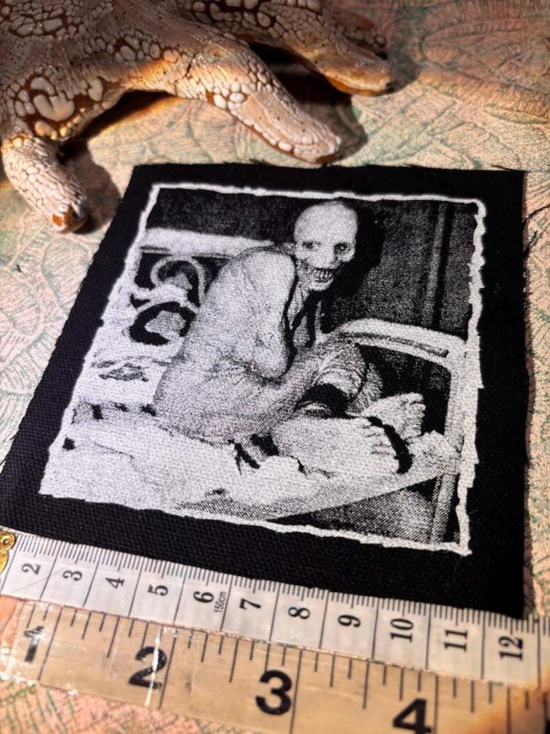 Puede incluir: Parche en blanco y negro con una imagen inquietante de una figura esquel&eacute;tica en una silla. El parche tiene un borde negro y un aspecto desgastado y texturizado. El parche mide aproximadamente 7,6 cm de ancho.