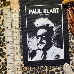 Peut inclure: Patch thermocollant noir et blanc représentant un portrait de Paul Blart, le flic du centre commercial, avec le texte "PAUL BLART MALL COP" au-dessus de l'image.