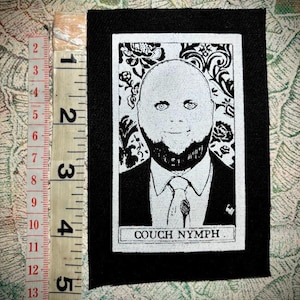 Pode incluir: Remendo preto e branco com um retrato de uma pessoa careca com barba, usando terno e gravata. O texto "COUCH NYMPH" está impresso na parte inferior. O remendo mede aproximadamente 12,7 cm de altura.