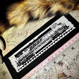 Puede incluir: Parche rectangular negro con una ilustración blanca detallada de un tren BNSF. El parche mide aproximadamente 15 cm de largo. Una cinta métrica se coloca en diagonal, mostrando medidas en cm. El fondo incluye tela estampada y piel.