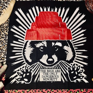 Könnte beinhalten: Schwarz-weiße Grafik eines Waschbären mit rotem Hut und weißen Strahlen. Der Text lautet "THE RICH ARE THE ONLY DANGEROUS MINORITY". Das Bild befindet sich auf einem schwarzen Hintergrund mit einem Leopardenmuster-Rand.