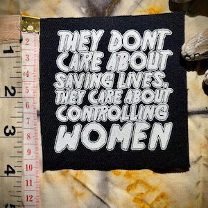 May include: Black square patch with white text: "THEY DONT CARE ABOUT SAVING LIVES, THEY CARE ABOUT CONTROLLING WOMEN." The patch is approximately 2 inches (5 cm) wide. The patch is on a patterned fabric.