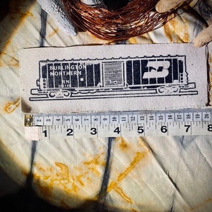 May include: A rectangular patch featuring a black and white illustration of a Burlington Northern train car. The patch is approximately 8 inches long. The text "BURLINGTON NORTHERN" and "BN 355145" are visible. The patch is on a textured fabric.
