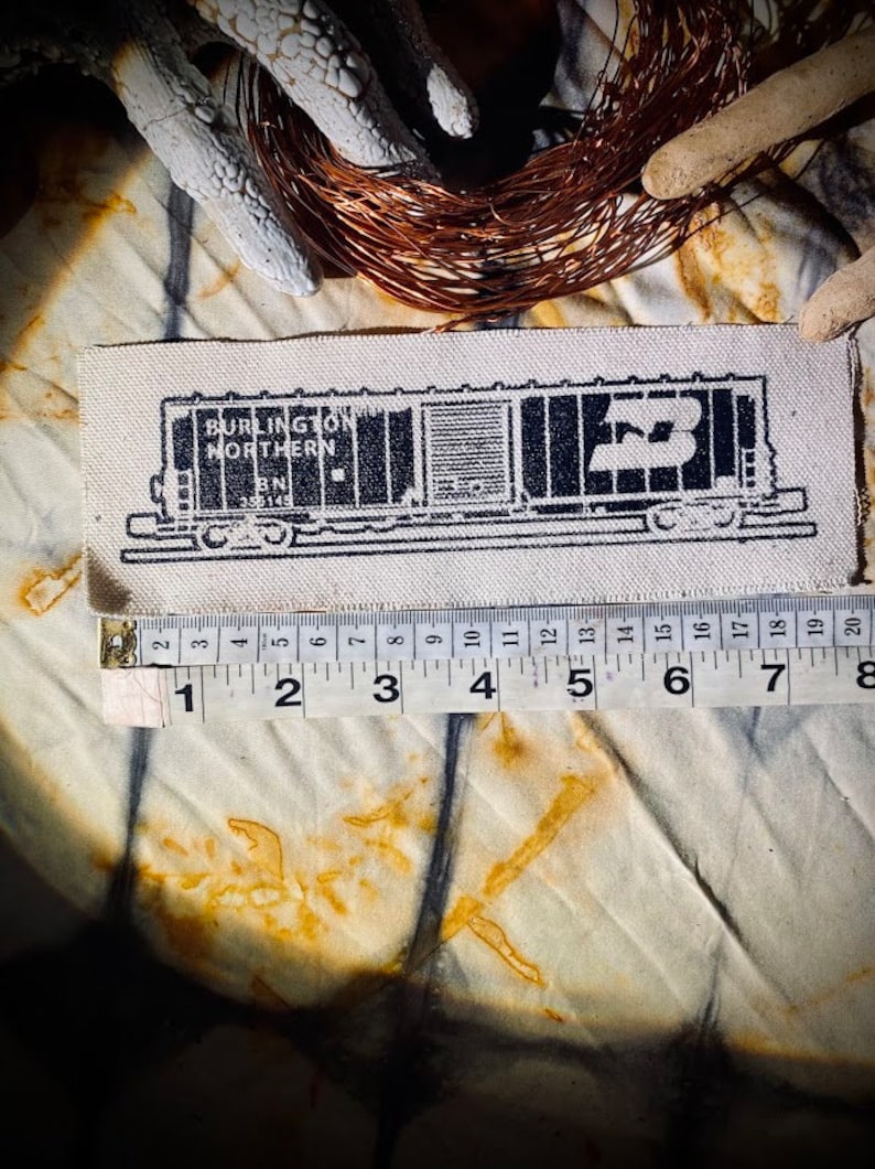 May include: A rectangular patch featuring a black and white illustration of a Burlington Northern train car. The patch is approximately 8 inches long. The text "BURLINGTON NORTHERN" and "BN 355145" are visible. The patch is on a textured fabric.