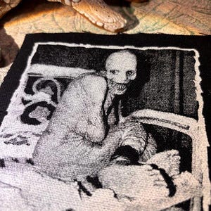 Puede incluir: Parche en blanco y negro que representa una figura esquel&eacute;tica con una amplia sonrisa, sentada en una silla. El parche tiene un borde negro y probablemente est&aacute; hecho de tela. Una serpiente bronceada y texturizada es parcialmente visible en el fondo.