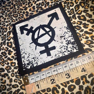 May include: A square black and white fabric patch featuring a central anarchy symbol combined with a transgender symbol. A measuring tape is positioned along the bottom edge, showing the patch width is approximately 4 inches across.