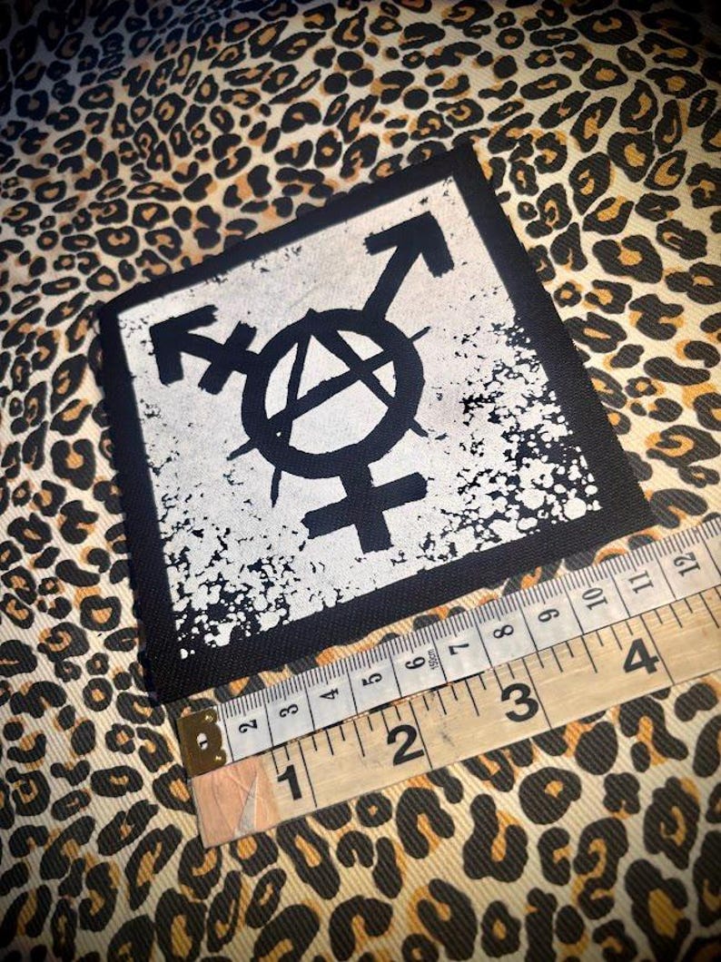 May include: A square black and white fabric patch featuring a central anarchy symbol combined with a transgender symbol. A measuring tape is positioned along the bottom edge, showing the patch width is approximately 4 inches across.