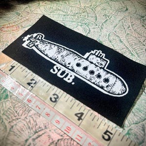 May include: A rectangular black fabric patch featuring a white screen-printed illustration of a submarine with the text SUB. positioned below it. A measuring tape is placed along the bottom edge, showing a length of approximately 11.5 centimetres.