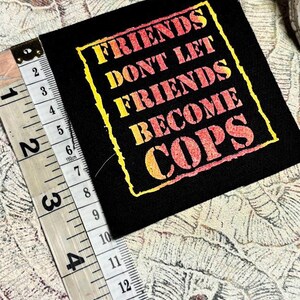 May include: A square black fabric patch featuring the yellow and orange gradient text "FRIENDS DONT LET FRIENDS BECOME COPS" inside a yellow border. A measuring tape placed along the left edge shows the patch is approximately 3.5 inches tall.