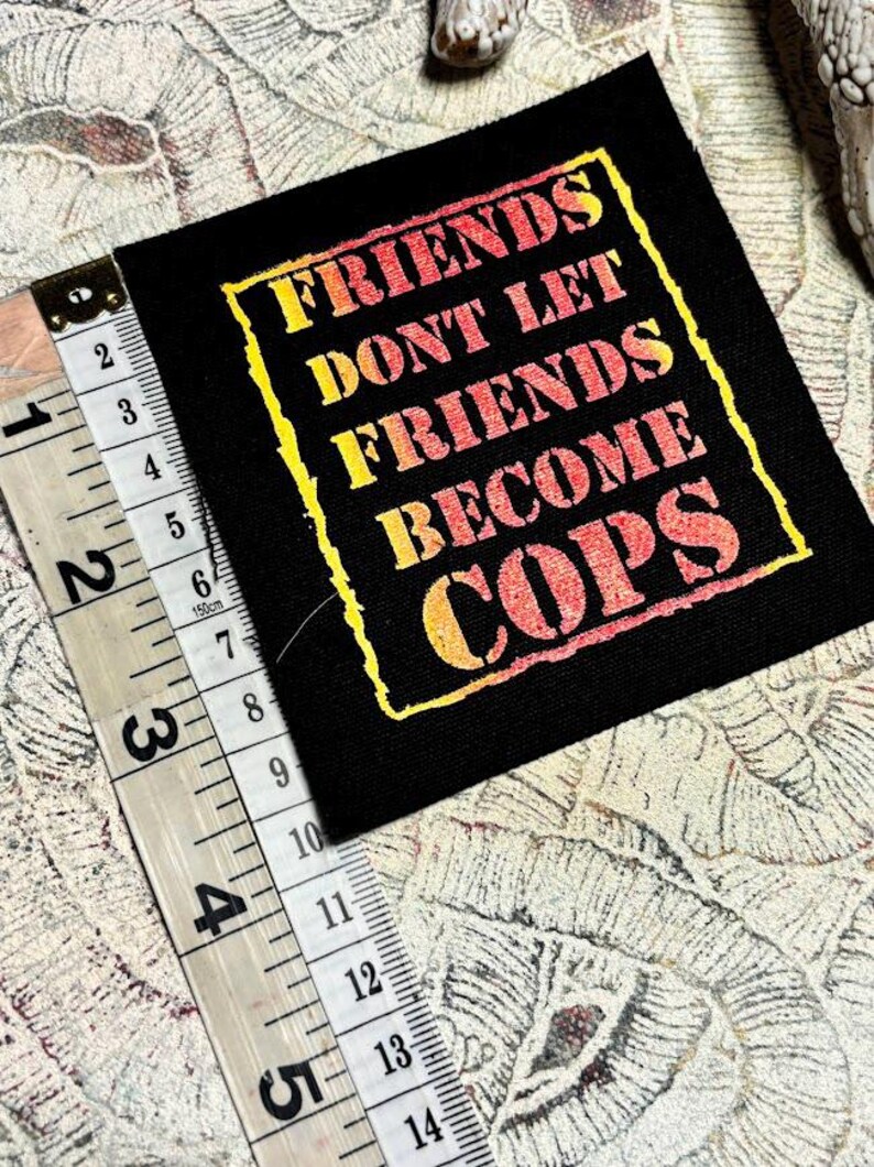 May include: A square black fabric patch featuring the yellow and orange gradient text "FRIENDS DONT LET FRIENDS BECOME COPS" inside a yellow border. A measuring tape placed along the left edge shows the patch is approximately 3.5 inches tall.