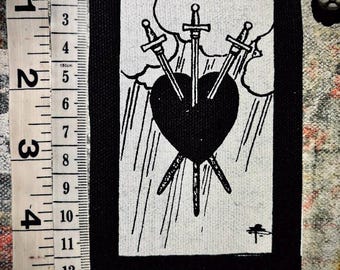 May include: A rectangular black and white fabric patch featuring a Three of Swords tarot card design with a heart pierced by three swords, positioned next to a measuring tape showing a length of approximately 5.5 inches and width of 3 inches.