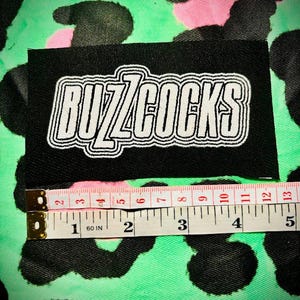 May include: Black patch with white text that reads "Buzzcocks" on a green and pink leopard print background.
