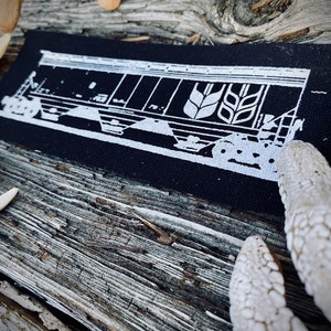 Op de afbeelding: Zwart-witte patch met een wit silhouet van een treinwagon met een tarwe-arenlogo aan de zijkant. De patch is gemaakt van zwarte stof met een witte rand.