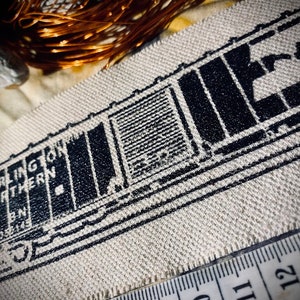 May include: Close-up of a beige fabric with a black train car design and the words "Burlington Northern". A measuring tape is along the bottom edge. Copper wire is in the background.