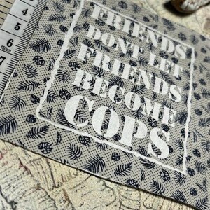May include: A square fabric patch with a light gray background featuring a dark blue repeating leaf pattern. The text FRIENDS DONT LET FRIENDS BECOME COPS is printed in white within a rough-edged white border. A measuring tape shows a width of 2.5 inches.