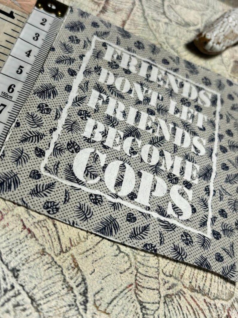 May include: A square fabric patch with a light gray background featuring a dark blue repeating leaf pattern. The text FRIENDS DONT LET FRIENDS BECOME COPS is printed in white within a rough-edged white border. A measuring tape shows a width of 2.5 inches.