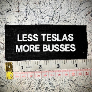 May include: Black fabric patch with white text that reads "LESS TESLAS MORE BUSSES". The patch is measured with a tape measure.
