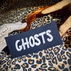 May include: A black fabric patch with white text that reads "GHOSTS" on a leopard print background.