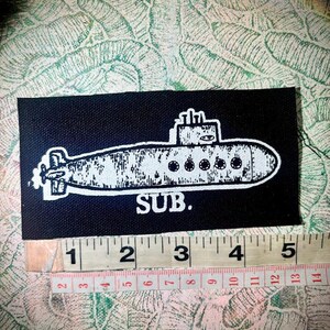 May include: A rectangular black fabric patch featuring a white screen-printed illustration of a submarine with the text SUB. below it. A measuring tape is positioned along the bottom edge, showing the patch is approximately 12.7 cm wide.