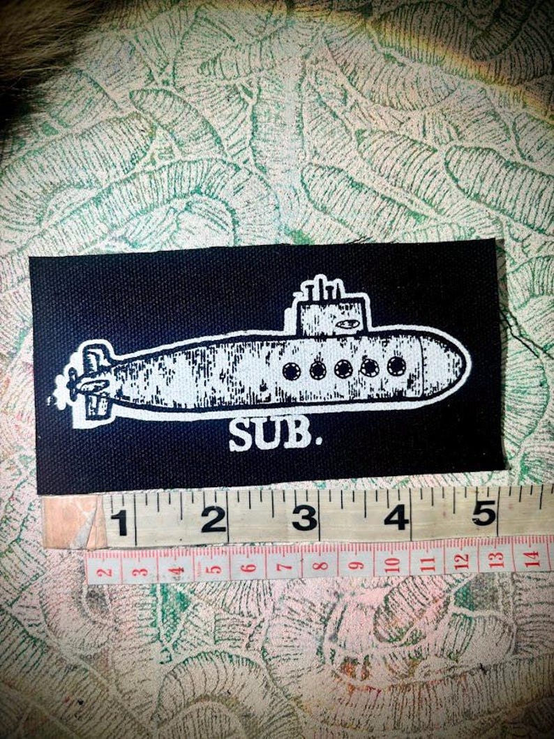 May include: A rectangular black fabric patch featuring a white screen-printed illustration of a submarine with the text SUB. below it. A measuring tape is positioned along the bottom edge, showing the patch is approximately 12.7 cm wide.