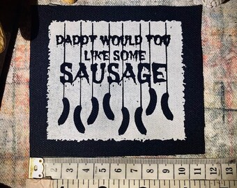 May include: A white patch with black text that reads "Daddy would you like some sausage" with a row of black sausage shapes hanging from the text. The patch is on a black background and is measured with a ruler.
