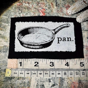 Può includere: Un'immagine in bianco e nero di una padella in ghisa con la parola "pan." stampata sotto. L'immagine è su uno sfondo bianco ed è circondata da un bordo nero.