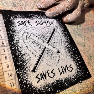 May include: Black and white patch featuring the text "SAFE SUPPLY SAVES LIVES" and a syringe design within a heart. The patch has a black border with white splatter. A measuring tape is visible. The patch is approximately 7.5 cm wide.