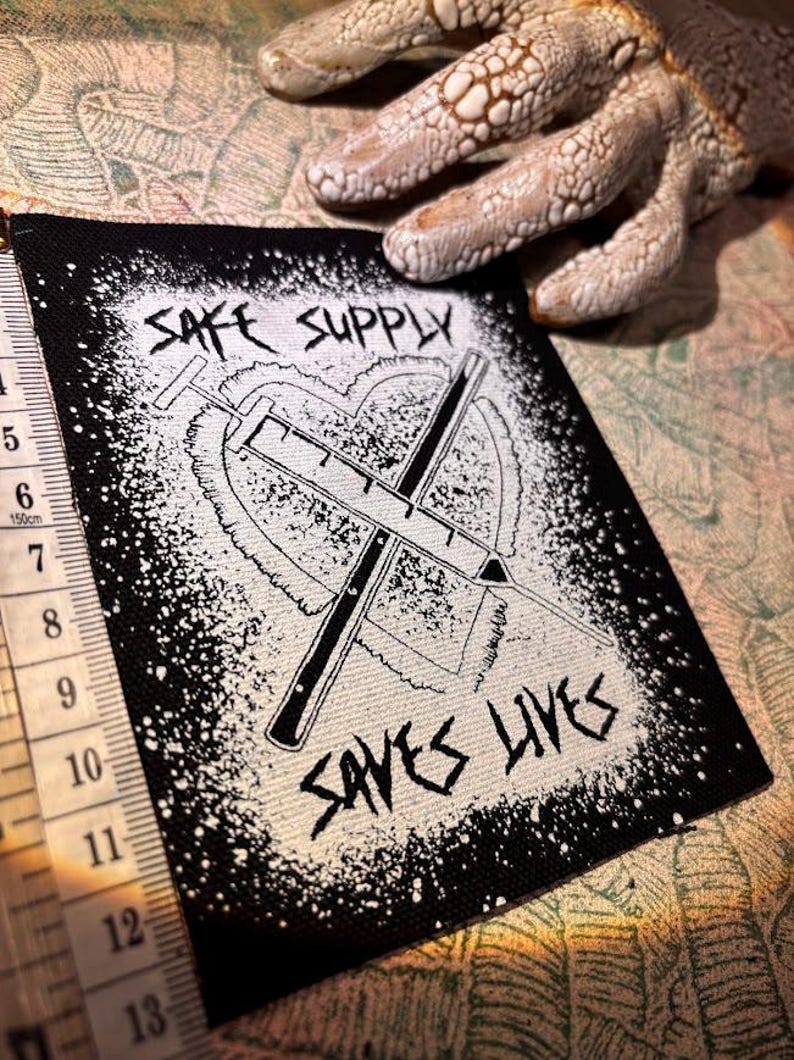 May include: Black and white patch featuring the text "SAFE SUPPLY SAVES LIVES" and a syringe design within a heart. The patch has a black border with white splatter. A measuring tape is visible. The patch is approximately 7.5 cm wide.
