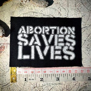 May include: A rectangular black fabric patch featuring white embroidered text that reads ABORTION SAVES LIVES. A measuring tape is positioned along the bottom edge, showing the patch width is approximately 11.5 centimetres across.