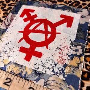 May include: A square fabric patch featuring a red anarchy symbol combined with a gender symbol printed on a white center square. The patch has a blue floral border and is positioned next to a ruler showing a width of approximately 2.5 inches.