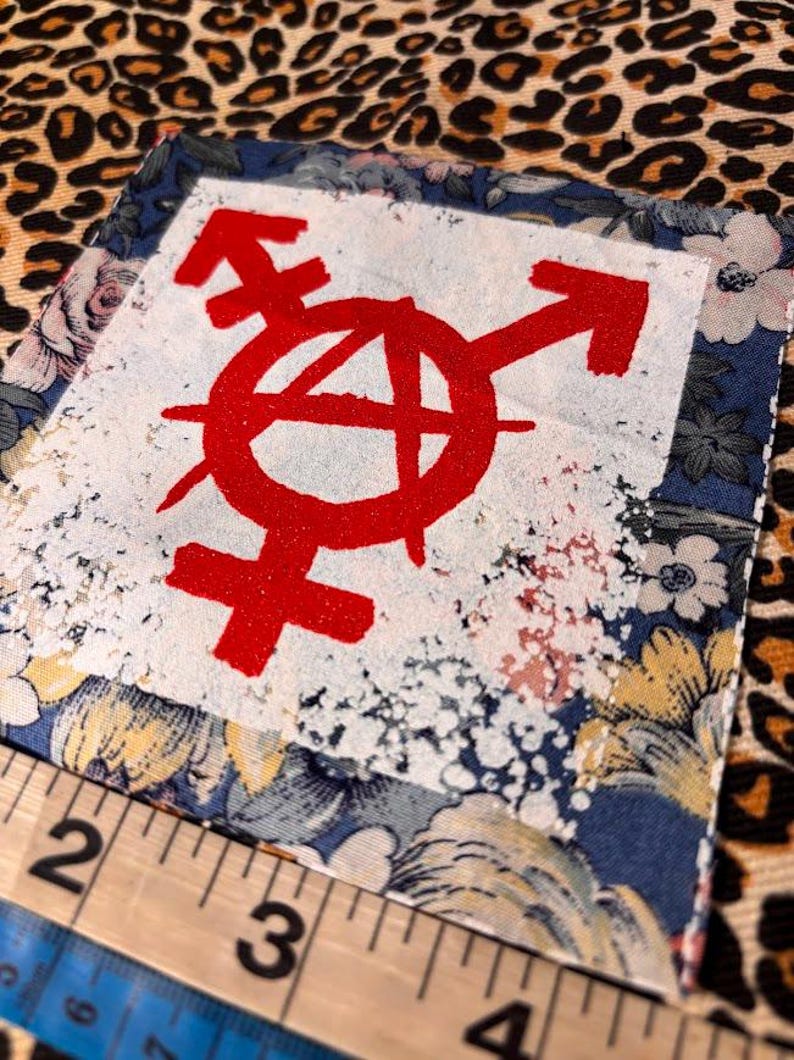 May include: A square fabric patch featuring a red anarchy symbol combined with a gender symbol printed on a white center square. The patch has a blue floral border and is positioned next to a ruler showing a width of approximately 2.5 inches.
