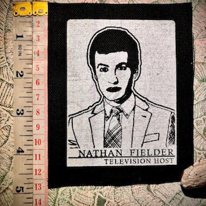 以下が含まれることがあります： テレビ司会者、ネイサン・フィールダーの肖像画が描かれた白黒のパッチ。画像は黒い縁で囲まれ、「NATHAN FIELDER TELEVISION HOST」の文字が含まれています。パッチの高さは約10cmです。