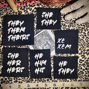 Peut inclure: Un ensemble de six patchs en tissu noir avec du texte blanc. Les patchs présentent différents ensembles de pronoms, notamment "They/Them/Theirs", "She/Her/Hers", "He/Him/His", "She/They", "He/They", et "Xe/Xem".