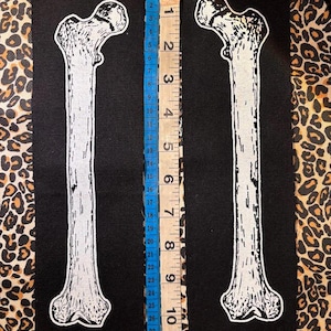 Pode incluir: Dois emblemas retangulares de tecido preto com uma estampa gráfica branca de um fémur humano. Uma fita métrica está colocada entre os emblemas, mostrando um comprimento de aproximadamente 30,5 cm para cada ilustração de osso no tecido.