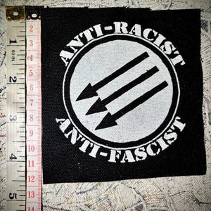 Könnte beinhalten: Schwarzer quadratischer Aufnäher mit weißem Text und Grafik. Der Text lautet "ANTI-RACIST" und "ANTI-FASCIST" um einen weißen Kreis mit drei nach unten zeigenden Pfeilen. Der Aufnäher misst ca. 10 cm x 10 cm.