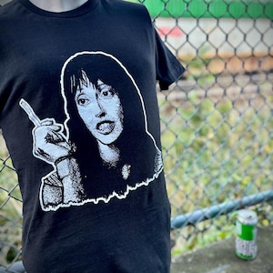 May include: Black t-shirt with a black and white graphic of a woman holding a weapon. The graphic is outlined in white. The t-shirt is displayed on a mannequin. A green and white can is in the background.