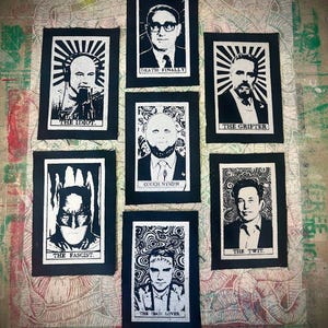 May include: A collection of seven black and white patches with portraits and text. The patches feature various individuals with text such as "THE IDIOT," "DEATH FINALLY," and "THE TWIT." The patches are arranged on a patterned surface.