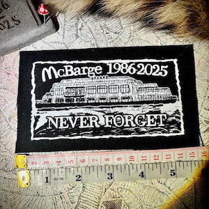 Puede incluir: Un parche rectangular negro con texto blanco y una ilustración detallada de un edificio. El texto dice "McBarge 1986-2025" y "NEVER FORGET". El parche mide aproximadamente 12,7 cm de largo.