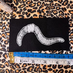 Op de afbeelding: Een rechthoekige zwarte stoffen patch met een witte lijntekening van een regenworm. Een meetlint is langs de onderrand geplaatst, waaruit blijkt dat de patch ongeveer 13 centimeter breed is.